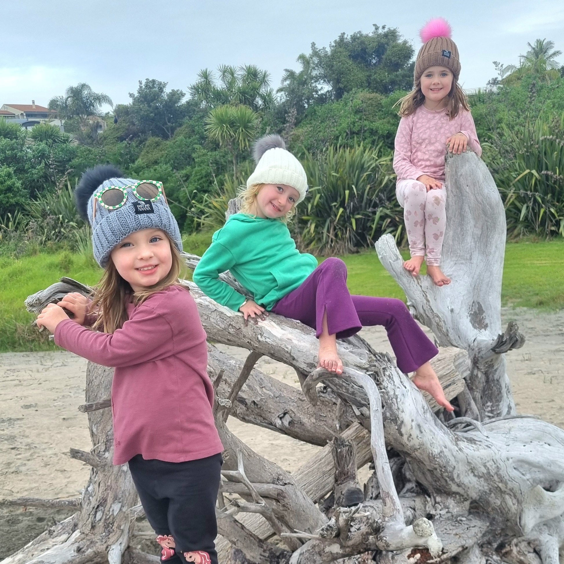 Three children playing on a large piece of driftwood at the beach.wearing Dolly & Naana beanies and sunglasses