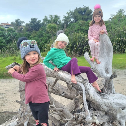 Three children playing on a large piece of driftwood at the beach.wearing Dolly & Naana beanies and sunglasses