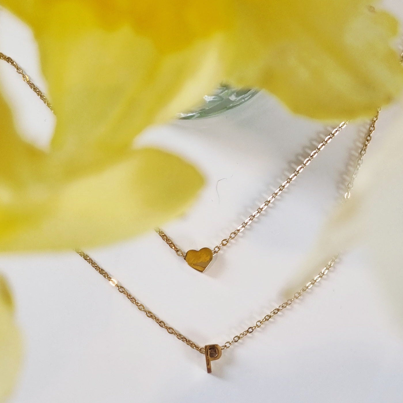 Two gold Love Letter Necklaces from Dolly & Naana with heart and letter p charm pendants on a white surface with yellow flowers in the background.