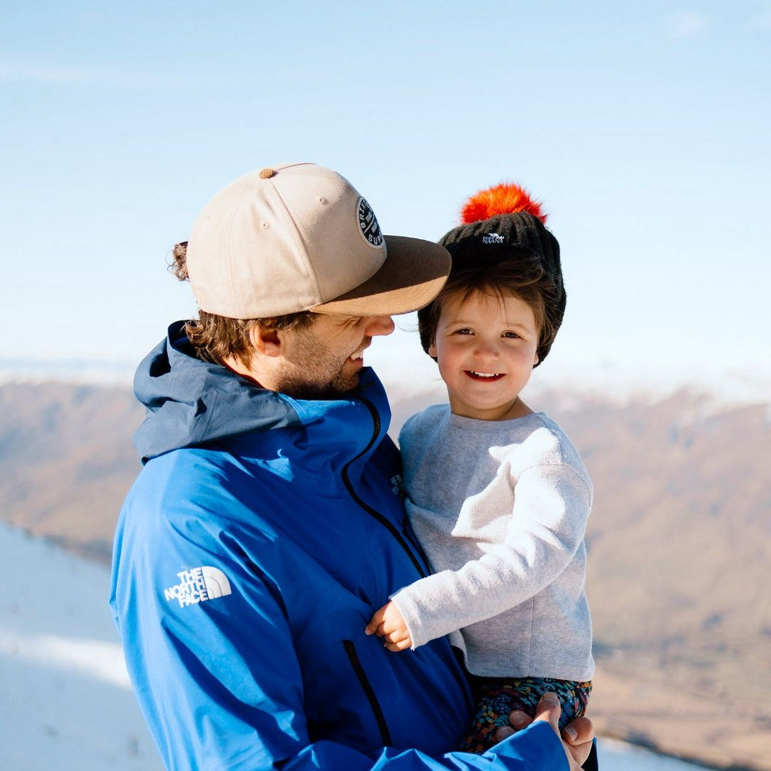 Person holding a child on a snowy mountain with a clear blue sky. wearing Dolly & Naana beanie