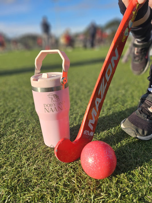 pink insulated stainless steel drink bottle for kids – Dolly & Naana with handle, red field hockey stick, and ball on grass