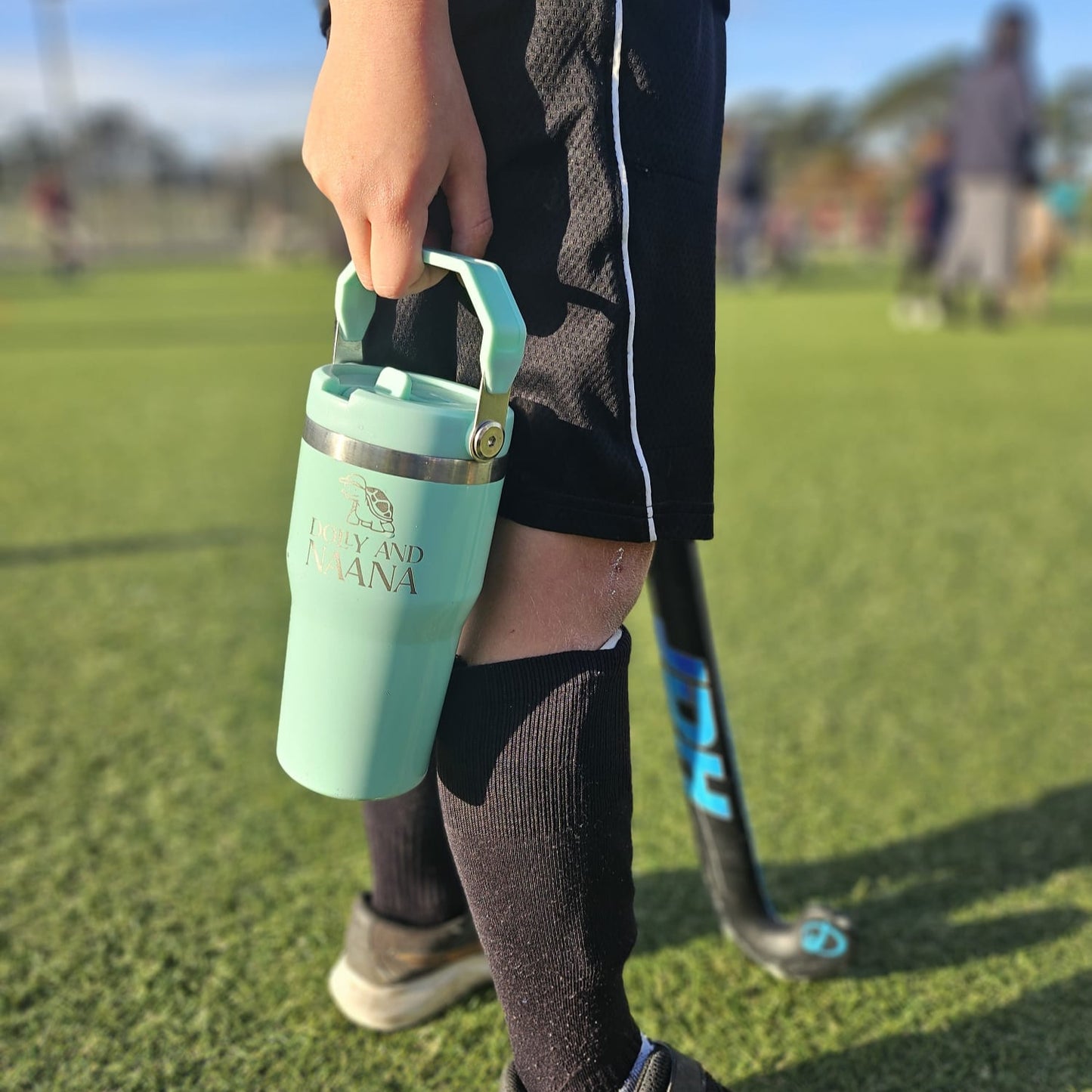 Person holding a Mint insulated stainless steel drink bottle for kids – Dolly & Naana with a sports field in the background