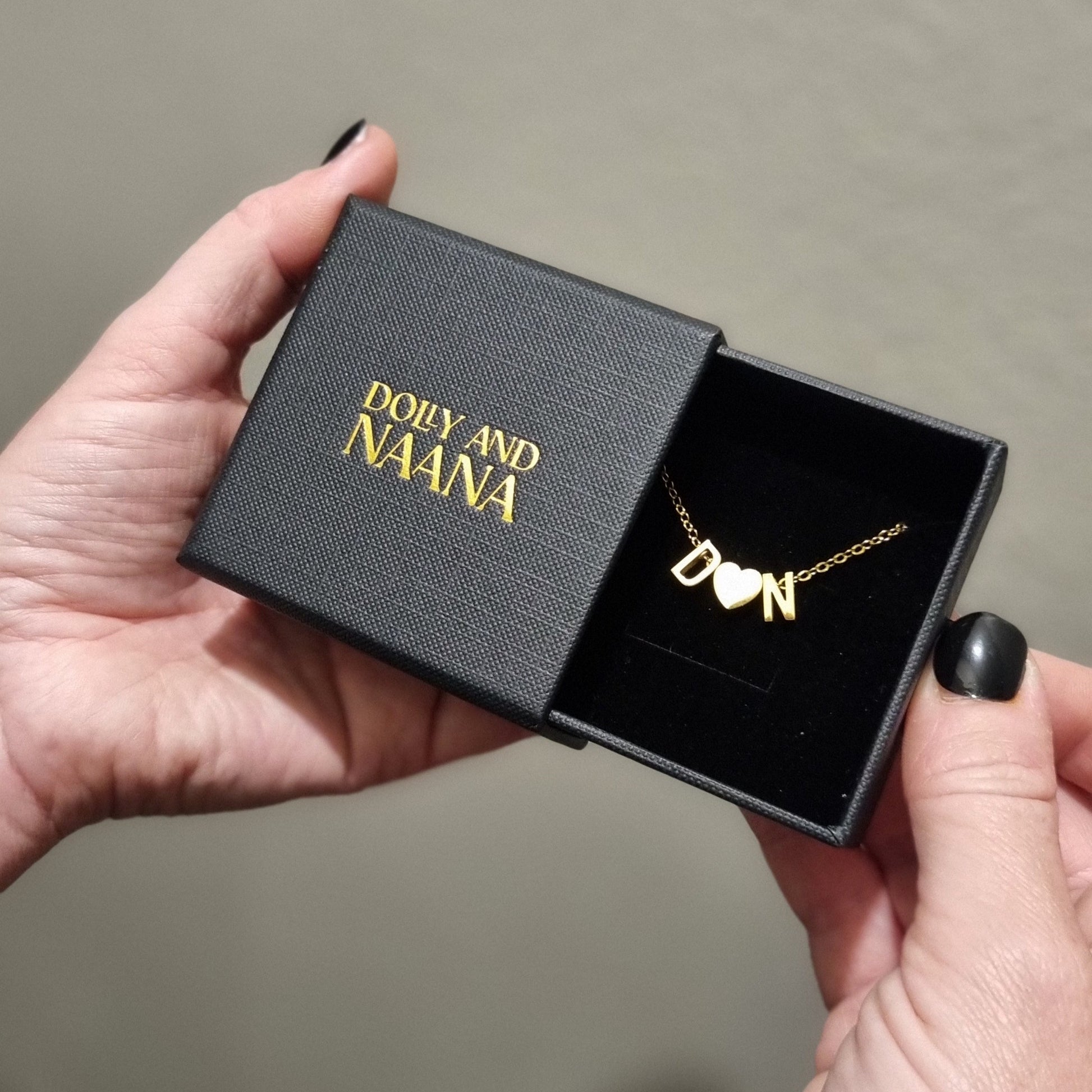Hand openinga black jewelry box with a gold Love Letter necklace inside from Dolly & Naana, against a neutral background.